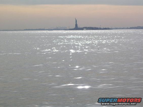 Water with sun glare, Statue of Liberty, and distant city skyline under overcast sky