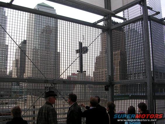 People observe a cross monument against a backdrop of skyscrapers, viewed through a metal fence