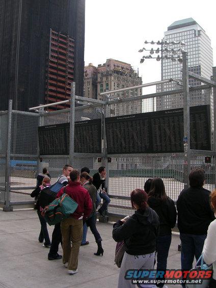 Memorial wall with names and people gathered around, tall buildings in background