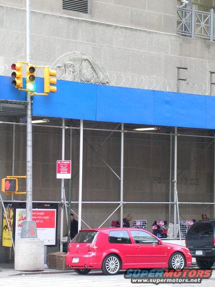 Red hatchback car parked on street with scaffolding and a post office sign in the background