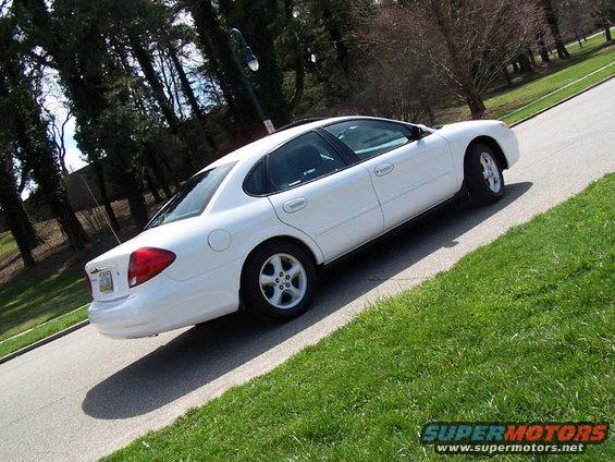 White sedan parked on a road with grass on the side. The car is viewed from the rear three-quarter angle