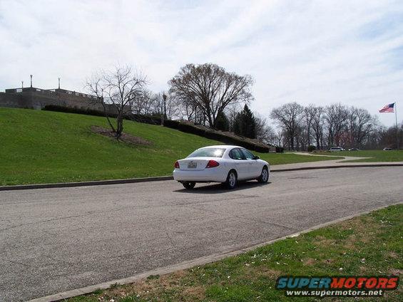 white sedan driving on a road with a grassy hill and trees in the background
