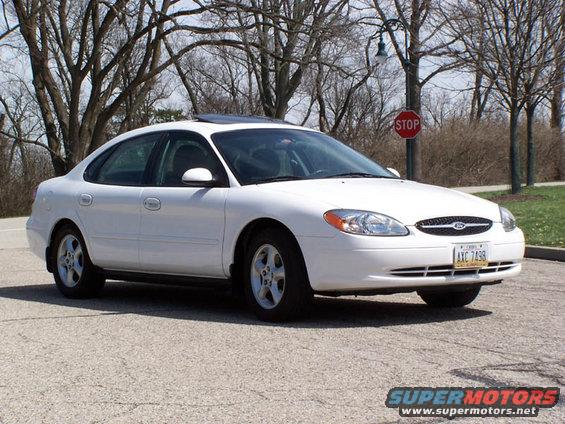 White sedan parked on asphalt, front quarter view. Visible license plate and stop sign