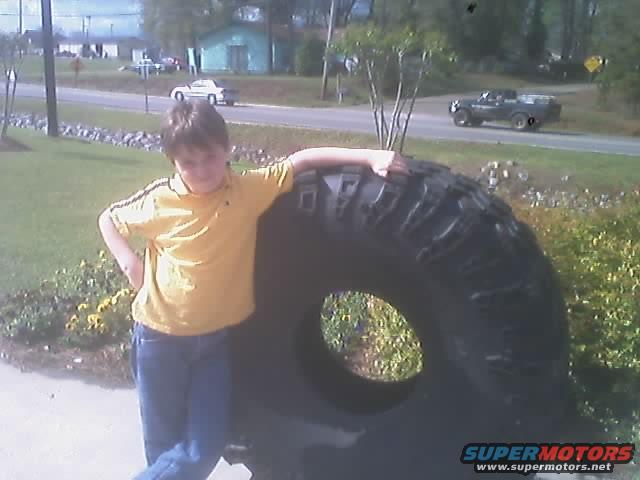 Boy in yellow shirt and blue jeans leaning on a large off-road tire