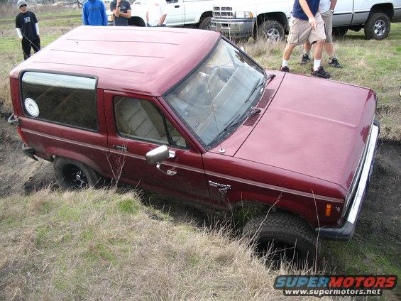 Red SUV stuck in a ditch with people watching. It looks like a Ford Bronco II