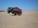 1985 Ford Bronco II - A red SUV is captured mid-air as it jumps over sand dunes under a clear blue sky.