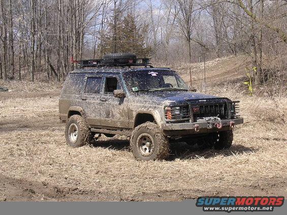 Muddy 1995 Jeep Cherokee with a roof rack and spare tire, parked in a wooded area
