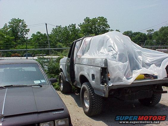 Gray Ford Bronco covered in plastic, parked next to a dark sedan