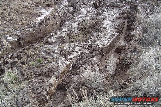 Muddy ground with deep tire tracks and dry brush. Supermotors.net logo visible