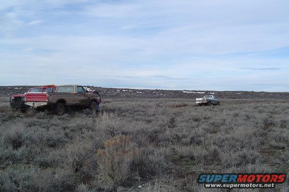 Brown Ford Bronco and red Ford truck parked in a field with sagebrush. Another truck is visible in the distance