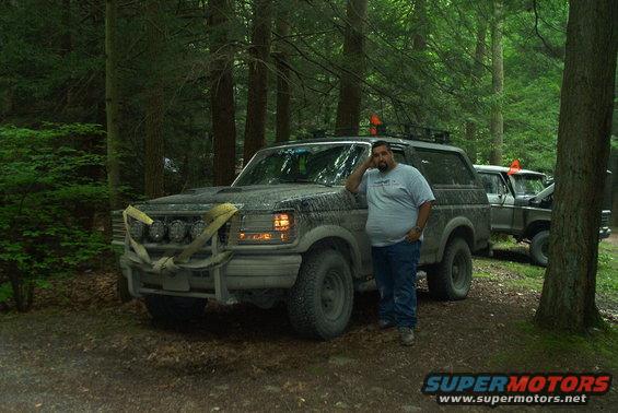 Mud-covered 1992 Ford Bronco with a bull bar and auxiliary lights, parked in a forest with a man leaning against it
