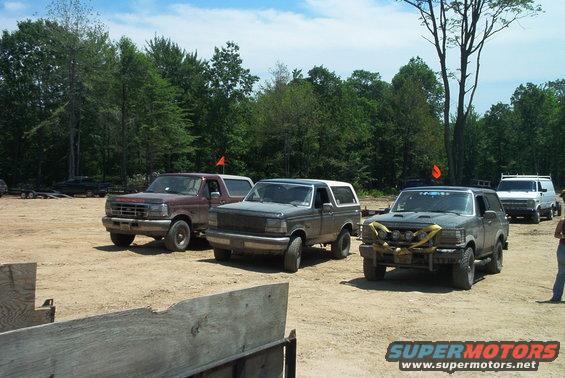 Three 1993 Ford Broncos parked in a dirt lot with trees in the background. A white van is visible behind the Broncos