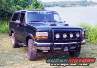 Dark green Ford Bronco with aftermarket grille guard and four fog lights, parked on grass near water
