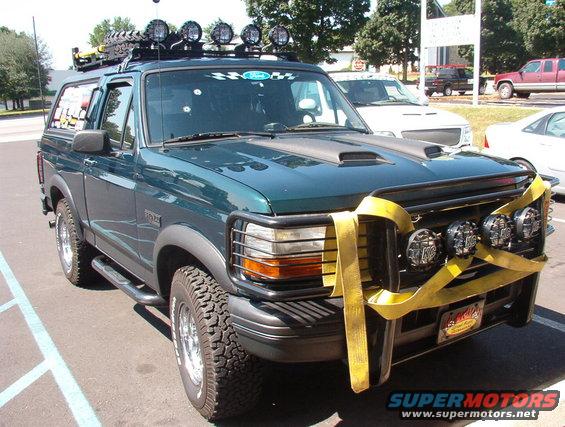 Green Ford Bronco with roof rack, off-road lights, and yellow straps on the grille