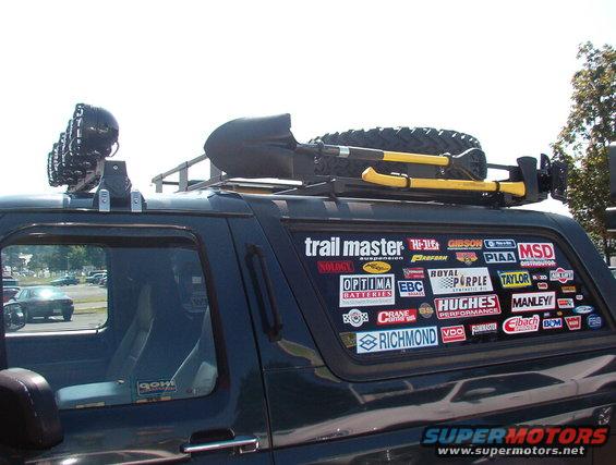 Side view of dark green SUV with spare tire, shovel, and axe on roof rack, covered in stickers