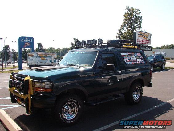 Dark green Ford Bronco with roof rack, spare tire, and many stickers on the rear window. Another SUV is parked nearby