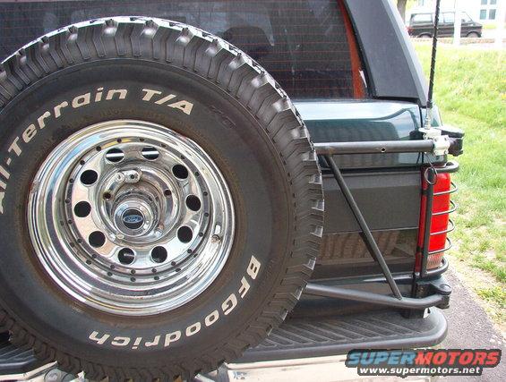 Spare tire mounted on the back of a 1993 Ford Bronco. Another vehicle is parked in the background