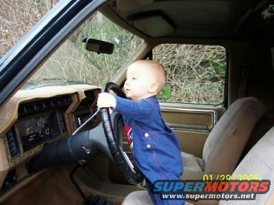 Baby wearing blue overalls grips steering wheel inside a tan interior of an older truck