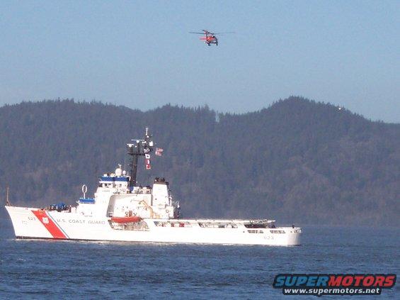 White U.S. Coast Guard cutter with red stripe, and a red helicopter in the sky