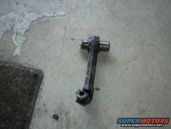metal automotive part with a pivot and a hole at the bottom, lying on a concrete floor