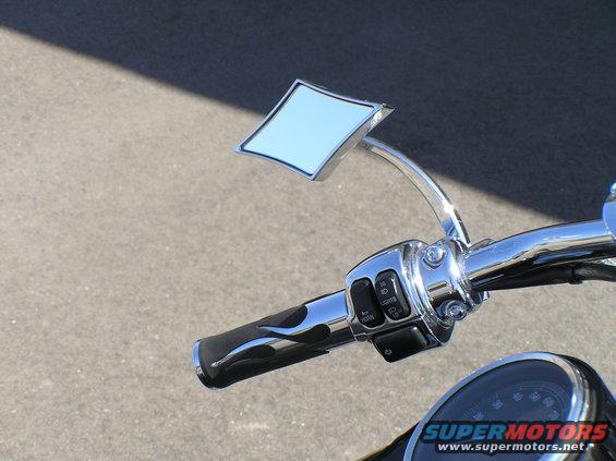 Motorcycle handlebars with flame grips, diamond-shaped mirror, and control buttons. Part of a speedometer is visible