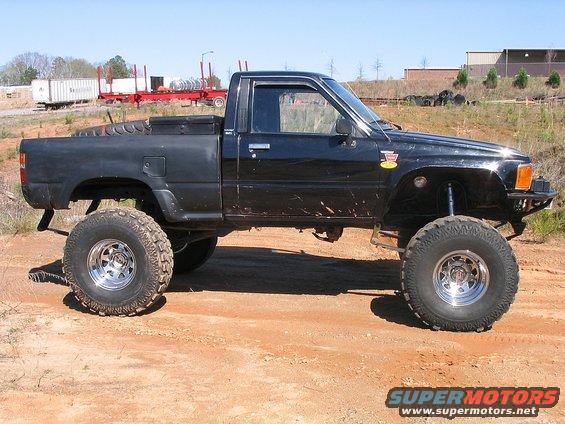 Lifted black pickup truck with large tires, parked on dirt. Has a toolbox in the bed