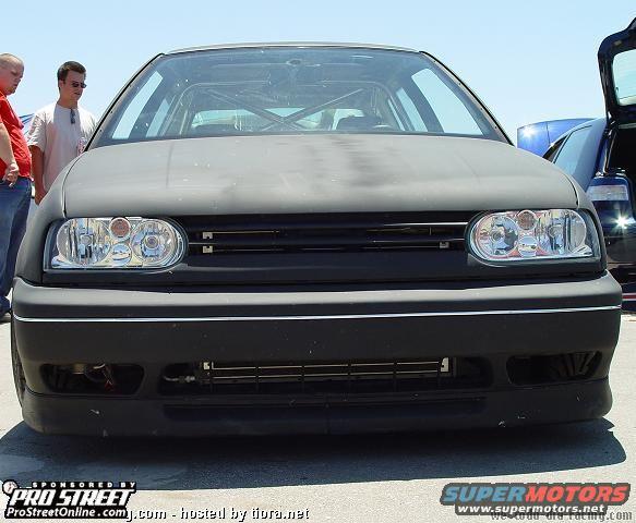 Front view of matte black Volkswagen Golf GTI with roll cage visible inside. Another car is parked behind it