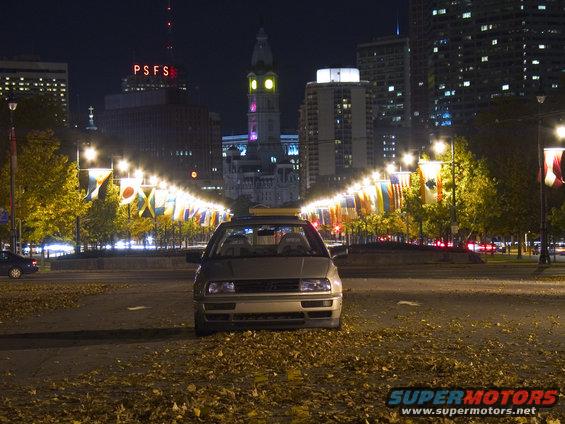 Silver sedan parked on a street at night, with city buildings and flags in the background