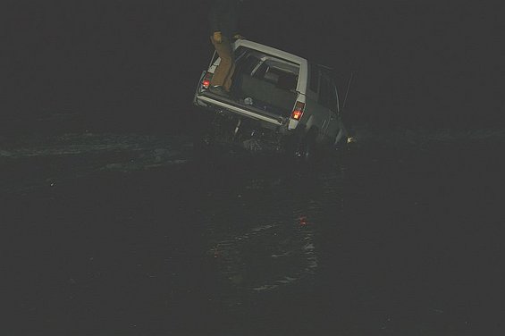 White SUV stuck in dark water, rear view with open trunk and person standing in it