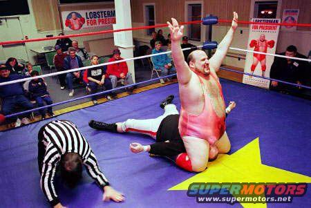 Wrestler in pink singlet on top of opponent, referee on mat, "Lobster for President" banner in background