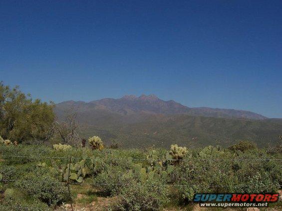 Desert landscape with cacti, scrub brush, and mountains under a clear blue sky. A "SUPERMOTORS" logo is visible in the lower right corner