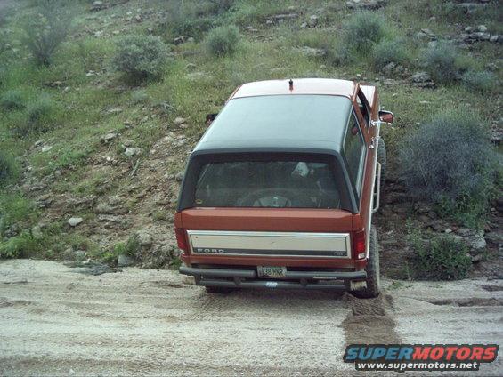 Rear view of an orange Ford Bronco with a black top driving on a dirt path in a hilly, brushy area. License plate reads J38 HNR