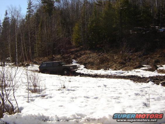 Dark SUV stuck in a muddy, snowy trail with trees in the background