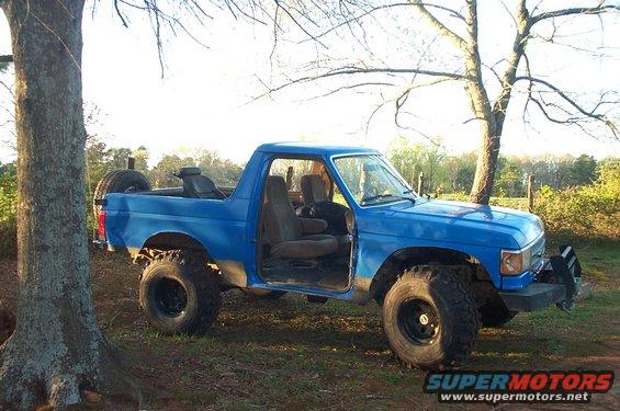 Blue Ford Bronco with lifted suspension and off-road tires, doors open, parked outdoors near trees