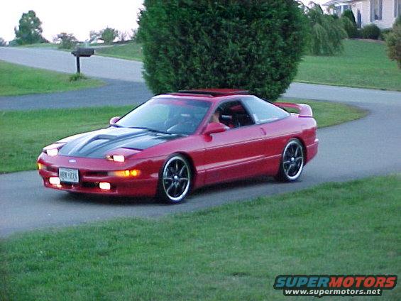 Red coupe with black hood and aftermarket wheels driving on a paved road