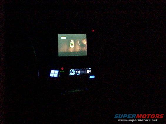Car interior at night, showing a small screen displaying a yellow car and a dashboard with glowing blue buttons