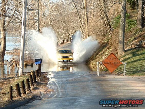 Yellow SUV driving through flooded road, creating large splashes. A "CLOSED ROAD FLOODED" sign is visible