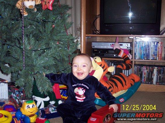Baby in a Santa outfit sits by a Christmas tree with toys. A TV and VCR are in the background