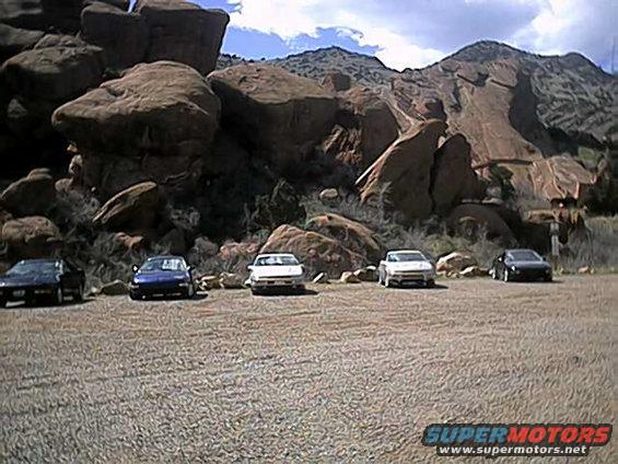 Five cars parked in a gravel lot with large rock formations and mountains in the background