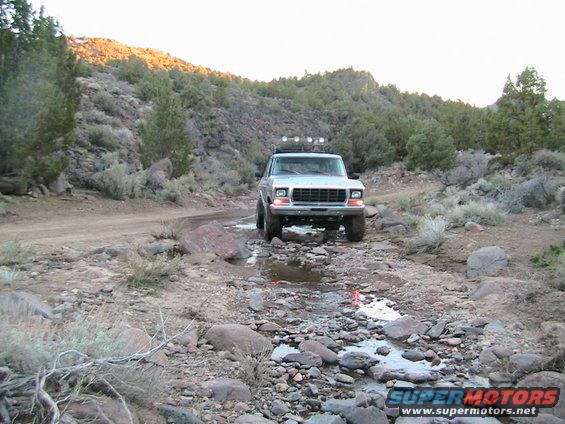 1978 Ford Bronco driving through a rocky stream bed with scrub brush and hills in background