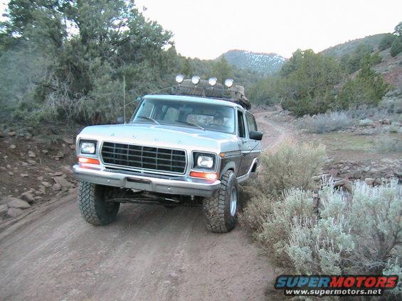 1978 Ford Bronco driving on a dusty, rocky trail with sparse vegetation and mountains in the background