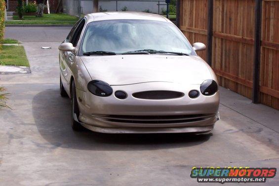 Tan sedan with blacked-out headlights and custom grille, front view