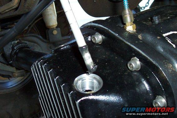 Close-up of a black differential housing with a tool inserting into a fill hole. A shock absorber is visible in the background