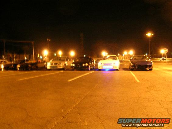 Several cars parked in a lot at night, one with blue underglow