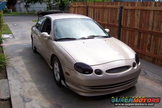 tan sedan parked in driveway, front end visible, wooden fence behind it