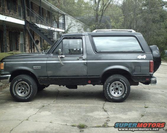 Gray 1987 Ford Bronco parked on concrete, side view, with a spare tire on the rear