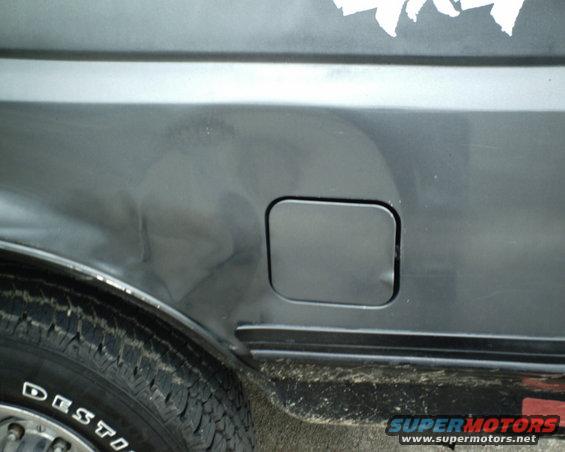 Close-up of a dented gray vehicle rear quarter panel with a fuel door and tire visible