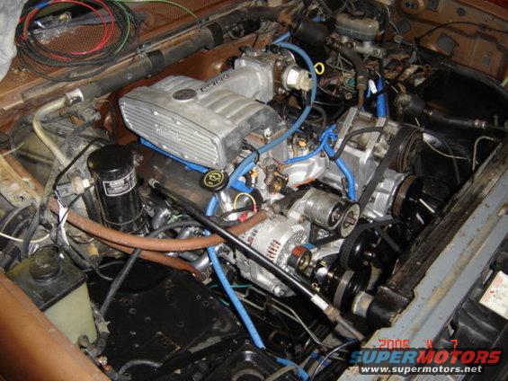 Engine bay with V8 engine, alternator, belts, and hoses. Water pump visible