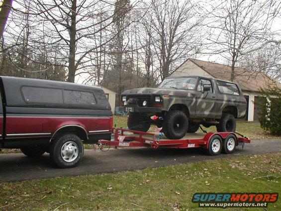 Camouflaged Ford Bronco on a red trailer being towed by a red pickup truck