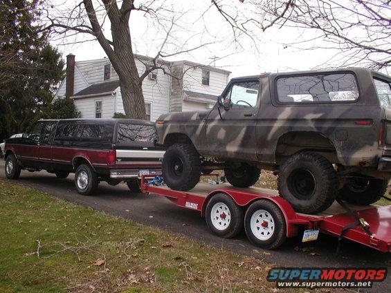 Camouflaged 1979 Ford Bronco on a trailer, towed by a pickup truck